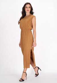 Abito pieghettato, di colore marrone uniforme, con un corpetto aderente e uno spacco laterale, realizzato in tessuto leggero. Abbinato a sandali neri con tacco alto.
