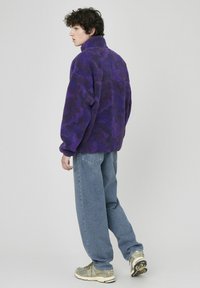 Veste en polaire violette avec un motif camouflage texturé, col haut et coupe décontractée, associée à un jean baggy bleu clair et des baskets.