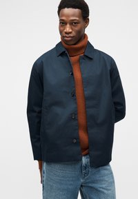 Μπλε ναυτικό μπουφάν με γιακά, με κούμπωμα με κουμπιά και πλαϊνές τσέπες, φορεμένο πάνω από ένα πουλόβερ με λαιμόκοψη σε χρώμα σκουριάς, συνδυασμένο με μπλε τζιν.