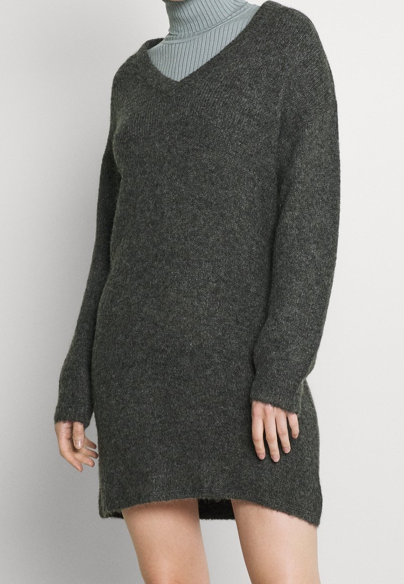 Femme portant une robe-pull oversize gris foncé à col en V, superposée à un col roulé côtelé gris clair, sur un fond uni.
