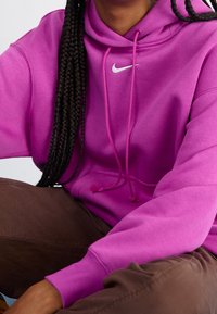 Pink Nike hoodie with a drawstring hood, features a white Nike logo on the chest, paired with brown pants. Soft fabric texture.