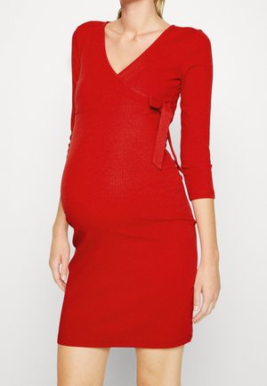 Femme enceinte portant une robe portefeuille rouge ajustée à manches trois quarts, debout devant un fond blanc uni.