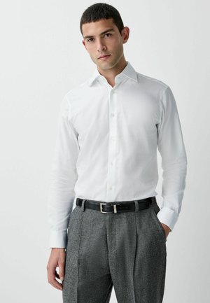 White long-sleeve shirt with a classic collar and buttons, paired with gray patterned trousers and a black belt with a metallic buckle.