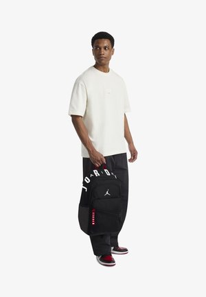 Zwarte Jordan-rugzak met wit logo, rode accenten en ritsdetails. Model draagt een off-white shirt en zwarte broek met een losse pasvorm.