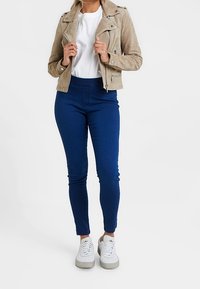 Vrouw draagt een beige suède motorjack, wit T-shirt, blauwe strakke jeans en witte sneakers, staand tegen een effen achtergrond.