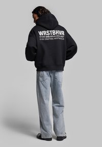 Felpa nera con cappuccio e testo bianco sulla schiena, abbinata a jeans in denim blu chiaro e scarpe nere; design relaxed fit.