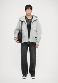 Sweat à fermeture éclair gris avec de grandes poches avant, porté sur un t-shirt noir, associé à un jean noir large et des baskets noir et blanc.