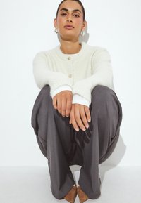 Šviesiai kreminė mezginė kardiganas su auksinėmis sagomis, derinamas su tamsiai pilkos spalvos plačiomis kelnėmis ir rudos spalvos smailių batų pora, rankos ilsisi ant kelių.