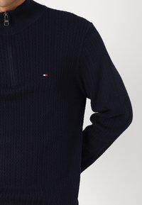 Pullover di maglia blu navy con colletto alto e chiusura con zip. Presenta un motivo a spina di pesce e un piccolo logo ricamato in rosso, bianco e blu.