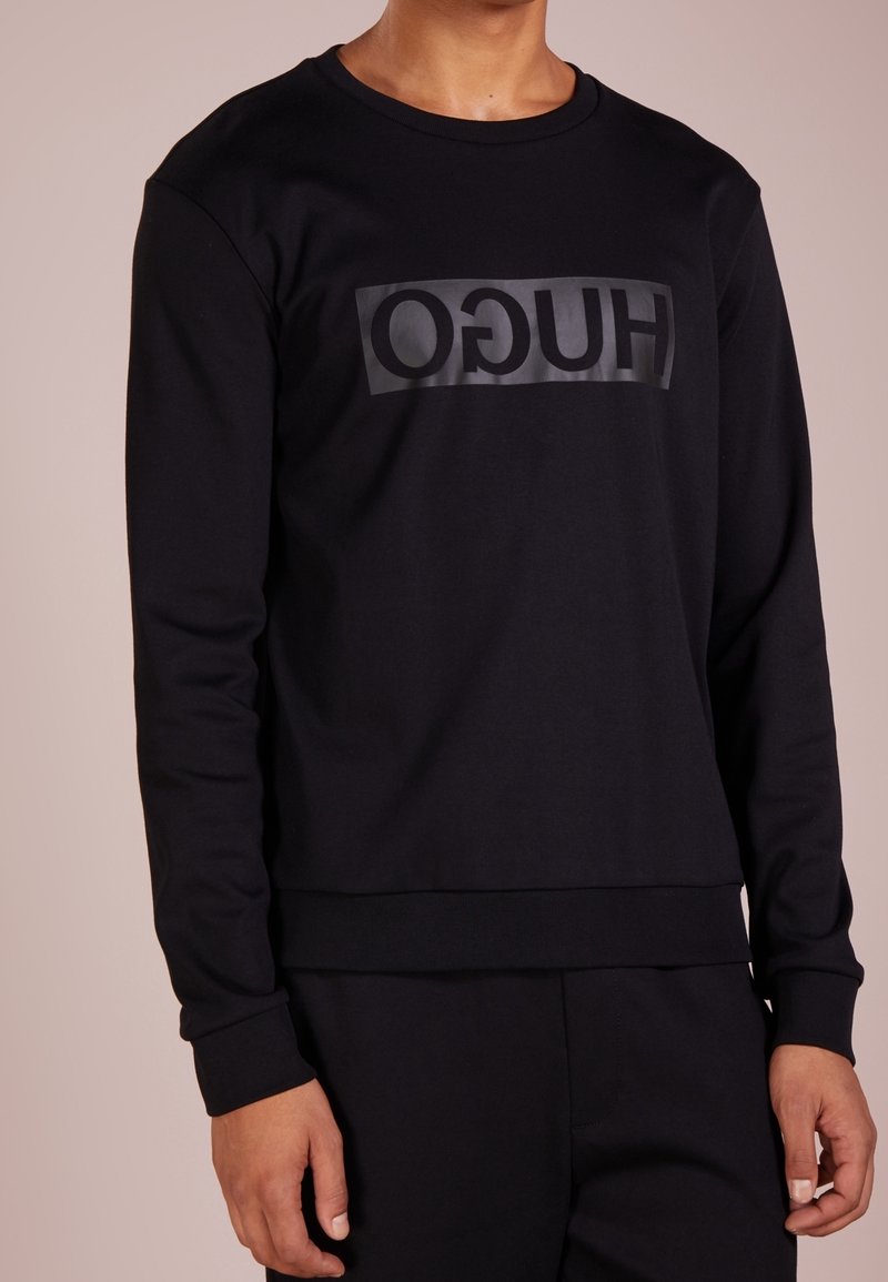 Schwarzer Hoodie aus Baumwolle, mit einem auffälligen, matten Logo auf der Brust in rechteckigem Design. Gerippte Bündchen und Saum.