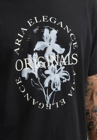 Sort skjorte med hvidt blomstermotiv og teksten "ORIGINALS" centreret, omgivet af cirkulær tekst med ordene "AFRIA ELEGANCE".
