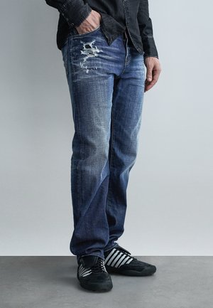 Άνδρας που φοράει ξεθωριασμένα μπλε σκισμένα τζιν, μαύρα και λευκά αθλητικά παπούτσια και μαύρο μακρυμάνικο πουκάμισο, με το ένα χέρι στην τσέπη, στέκεται σε γκρι πάτωμα.