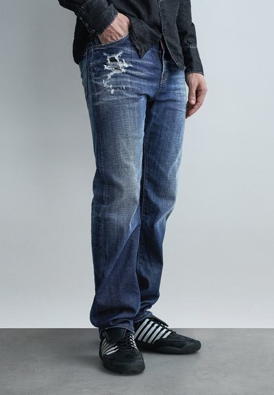 Homme portant un jean bleu délavé déchiré, des baskets noires et blanches, et une chemise noire à manches longues, avec une main dans la poche, debout sur un sol gris.