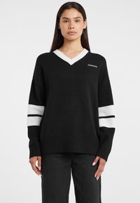 Pull noir à col en V avec col côtelé blanc et rayures blanches sur les manches. Texture tricotée, coupe décontractée, avec un petit logo "GUESS" sur la poitrine.