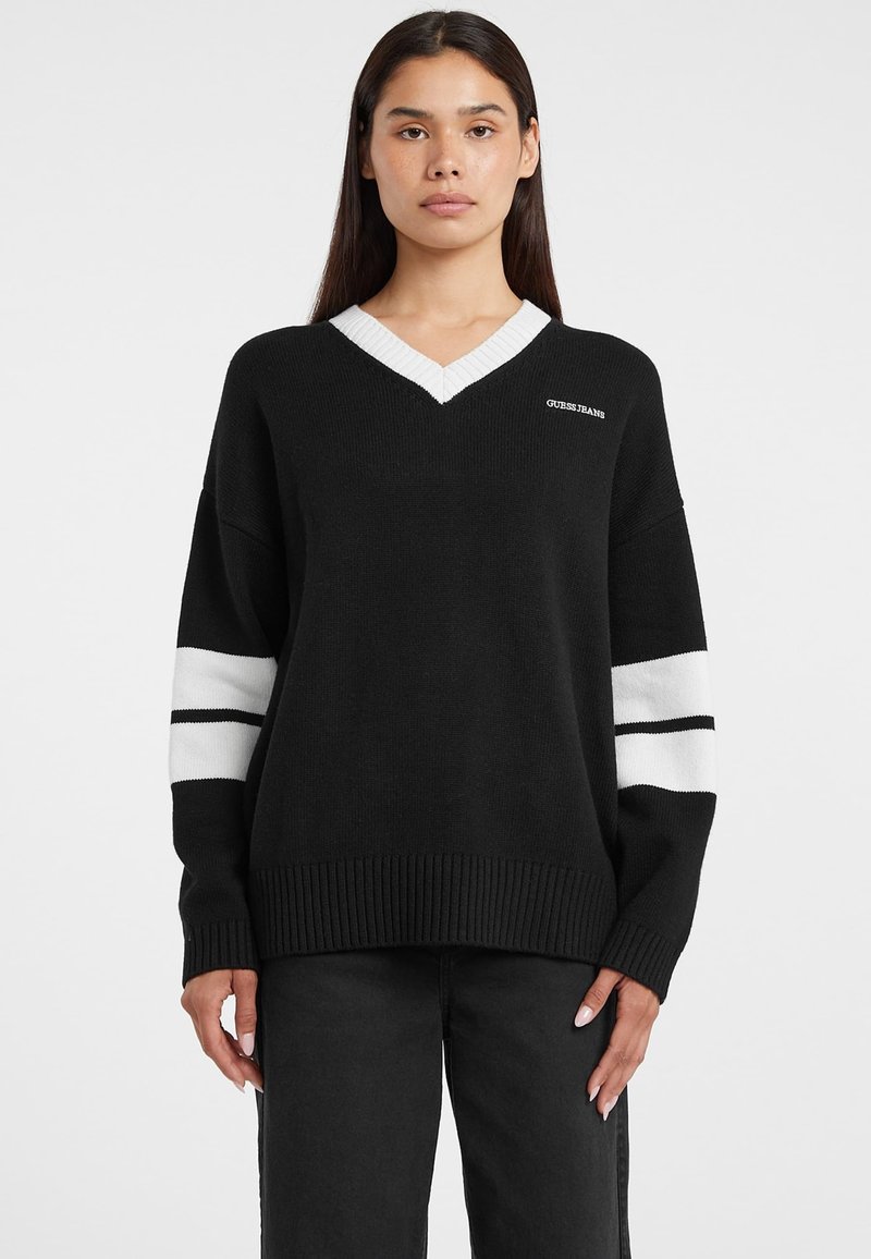 Pull noir à col en V avec col côtelé blanc et rayures blanches sur les manches. Texture tricotée, coupe décontractée, avec un petit logo "GUESS" sur la poitrine.