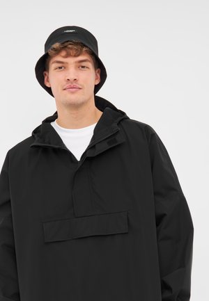 Chaqueta de pullover negra impermeable con un alto cuello y bolsillo frontal. Combinada con un sombrero de cubo negro a juego. Textura suave, diseño minimalista.