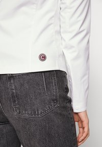 Gros plan sur une personne portant une veste blanche et un jean noir, montrant le bas de la veste avec un patch rond logo.