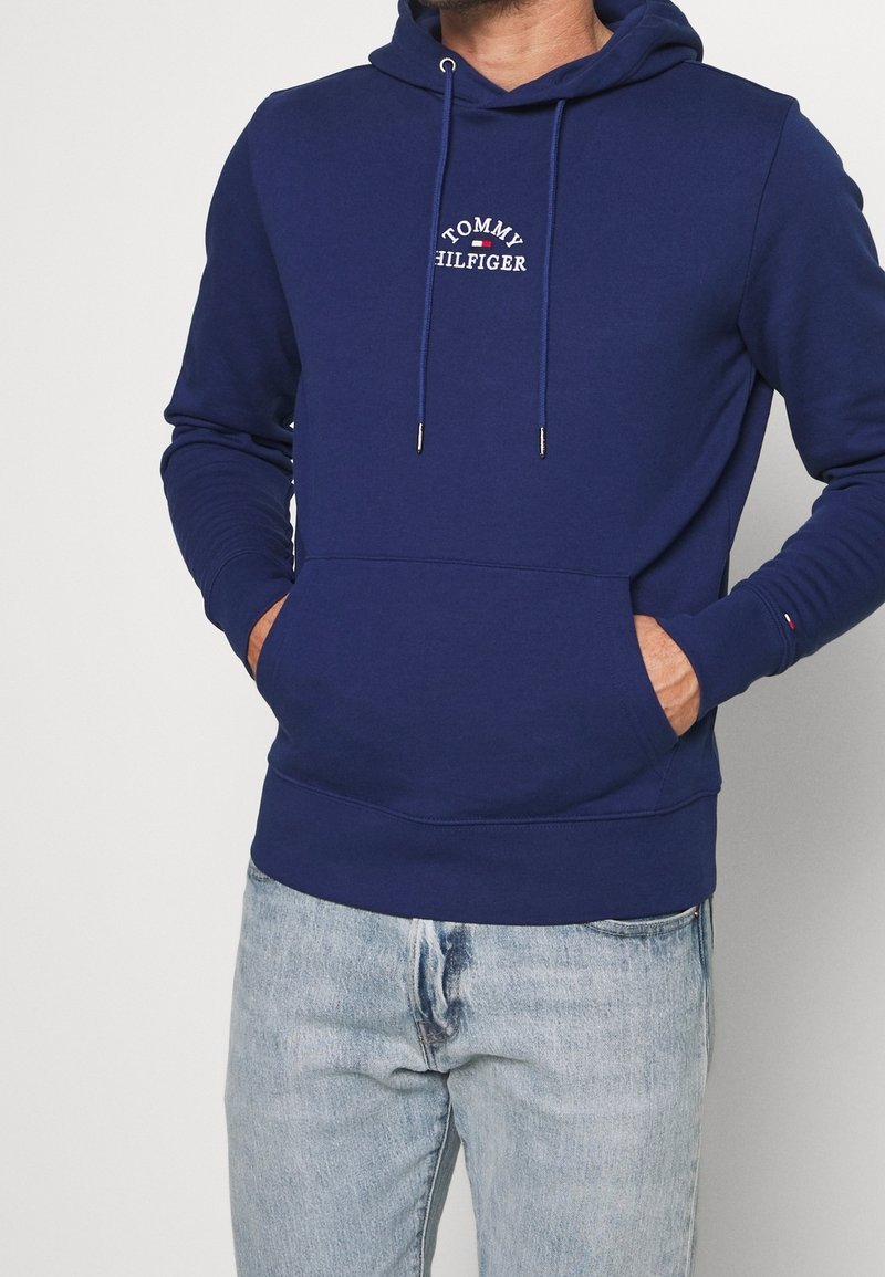 Homme portant un sweat à capuche Tommy Hilfiger bleu marine avec poche avant et logo, associé à un jean en denim bleu clair, mains dans les poches.