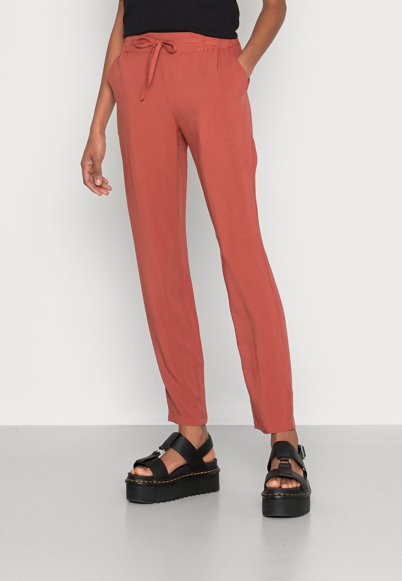 Persoon in roestrode drawstring broek en zwarte platform sandalen, staand tegen een effen lichtgrijze muur en vloer.