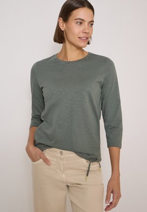 Femme aux cheveux courts bruns portant un haut vert à manches longues et un pantalon beige, debout avec une main dans la poche devant un fond uni.