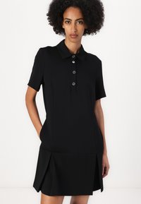 Abito polo nero con maniche corte, pattina con bottoni, tasche laterali e orlo plissettato. Realizzato in tessuto liscio e elastico.