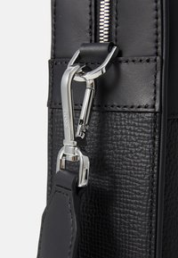 Détail d'un sac en cuir noir texturé avec un clip en métal argenté et une fermeture éclair, accentué par des éléments en cuir lisse contrastant.