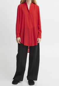 Maglietta rossa a maniche lunghe con colletto e patta di bottoni, abbinata a pantaloni neri a gamba larga e scarpe nere slip-on. Tessuto dalla superficie liscia.