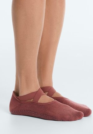 Legs wearing rust-colored ballet-style slip-on socks with crisscross elastic straps and textured grip soles on a smooth surface.