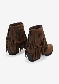 Brown suede ankle boots with fringe detailing, side zippers, and a low, solid black heel. Smooth texture and curved shape.