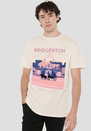 Junger Mann trägt ein cremefarbenes Bridgerton-T-Shirt mit Illustration einer Pferdekutsche und einem Herrenhaus in London, steht mit der Hand in der Tasche.