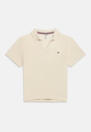 Polo de manga corta beige con cuello abierto y pequeño logo bordado en rojo, blanco y azul en el pecho izquierdo.