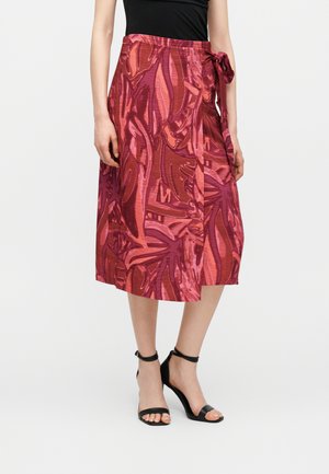 Kvinde iført et knælængde, rød og lyserød abstrakt mønstret omslagsnederdel bundet i siden, sort top og sorte ankelremssandaler.