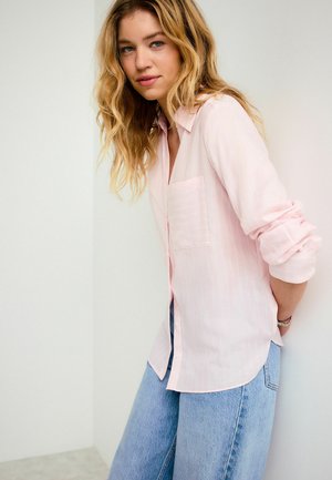 Młoda kobieta o falowanych blond włosach, ubrana w jasnoróżową koszulę na guziki i jasnoniebieskie jeansy, opiera się o białą ścianę.