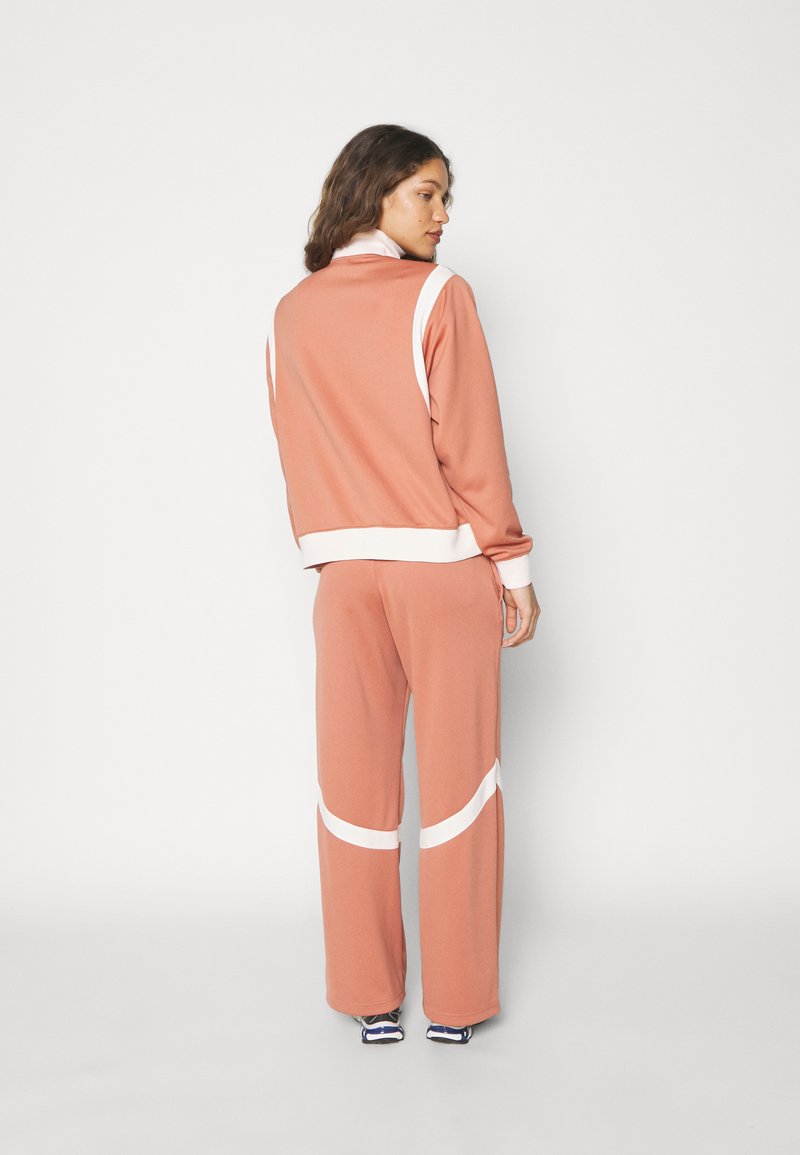 Ensemble de survêtement rose avec des accents blancs ; le haut présente un col montant et des poignets côtelés, tandis que le pantalon est ample avec des détails arrondis.
