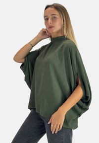 Grøn batwing-top med høj krave og korte delte ærmer, lavet af tekstureret stof. Stylet med mørkegrå jeans.