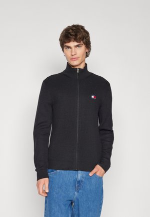 Pull à zip noir côtelé avec un col montant, présentant un petit logo tricolore sur la poitrine et une coupe ajustée.