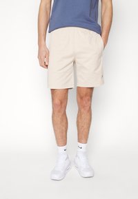 Beige cotton blend shorts with an elastic waistband, side pockets, and a small logo detail. Worn with a blue top and white sneakers.