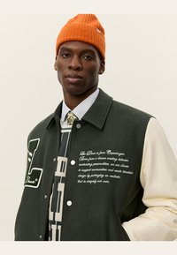 Grøn jakke i uldblanding med cremefarvede ærmer, prydet med kontrasterende broderi, et "L" logo og trykknapper. Modellen bærer en orange strikket beanie.