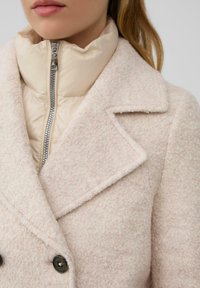 Frau in einem geschichteten, hellbeigen, strukturierten Wollmantel über einer cremefarbenen, gesteppten Jacke mit silbernem Reißverschluss, die den Kragen- und Knopfdetails zeigt.