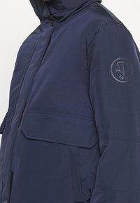 Mörkblå vinterjacka med hög krage, dragkedja och ficka med lock, som har ett emblem med skala och svärd på den övre vänstra ärmen med året 1855.