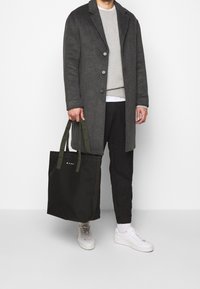 Cappotto di lana grigio, felpa grigio chiaro, pantaloni neri e sneakers bianche. Borsa a tracolla in tessuto nero con manici verdi olive e dettaglio del logo.