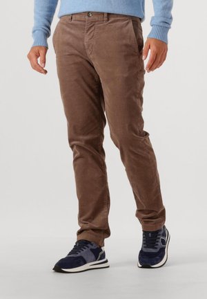 Mann in braunen Cordhosen, hellblauem Pullover und marineblauen Turnschuhen, der vor einem schlichten weißen Hintergrund steht.