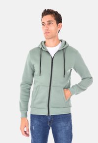 Felpa verde chiaro con zip, cappuccio regolabile, tasca a marsupio e zip nera. Abbinata a jeans blu strappati.