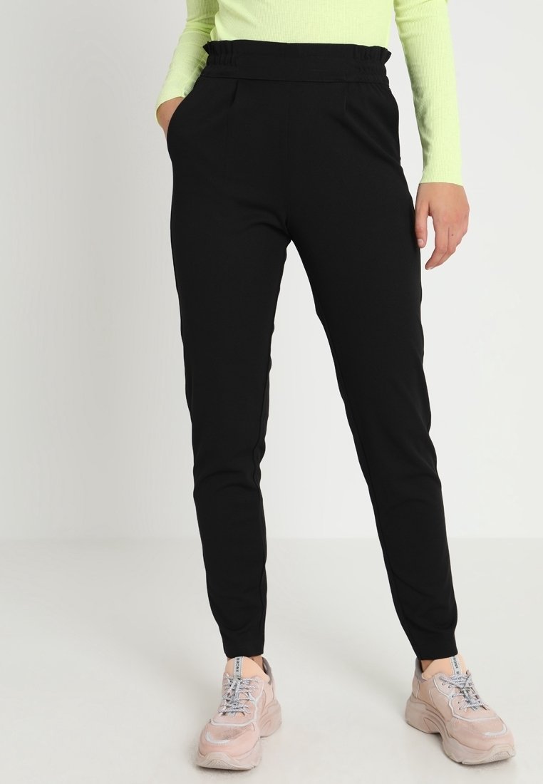 Pantalon de survêtement noir, effilé, avec une taille élastique et des poches latérales. Porté avec des baskets rose clair, dotées de lacets et d'un rembourrage.