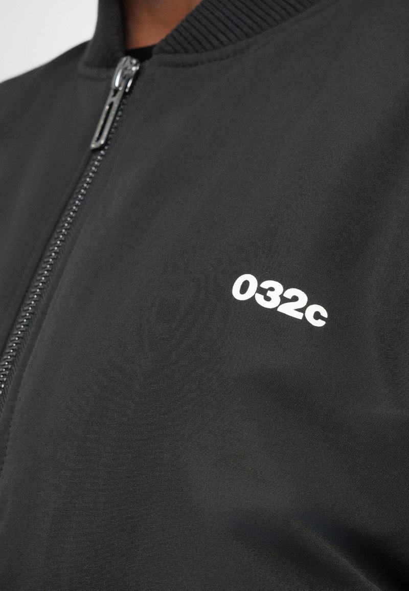 Veste bomber noire avec fermeture éclair, col côtelé et un petit logo blanc "032c" sur la poitrine. Texture de tissu lisse.