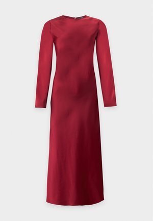 Femme aux longs cheveux blonds portant une robe rouge à manches longues, longueur genou, et des chaussures noires à talons, debout devant un fond clair uni.