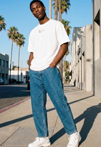 Hombre con camiseta blanca y vaqueros azules de pie en una calle soleada con palmeras y edificios al fondo.