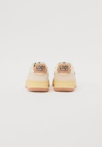 Sneakers Lacoste mają kremową cholewkę z zielonymi akcentami, brązowy naszywkę na pięcie oraz teksturowaną pomarańczową gumową podeszwę. Oznaczone brandingiem "L001".