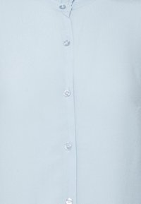 Camicia azzurro chiaro con una texture morbida, dotata di colletto a punte e piccoli bottoni rotondi traslucidi lungo il davanti.