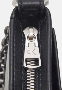 Černá kožená kabelka na zip se stříbrnými kovy, obsahující logo štítek a strukturovaný povrch. Na jedné straně jsou viditelné řetízkové ozdoby.